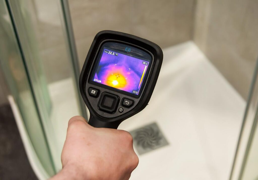 Hand holding a thermal imaging camera displaying a heat map over a shower tray.