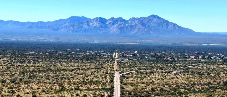A road stretches through a vast desert landscape toward a distant town beneath a range of large, rugged mountains.