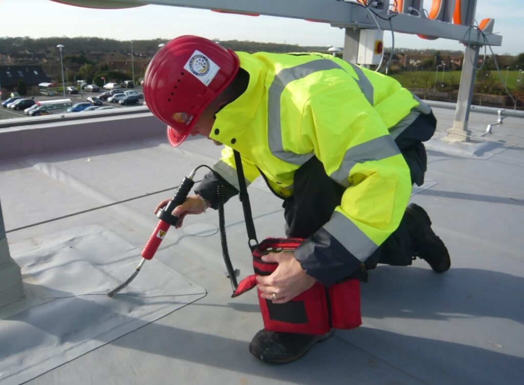 Construction worker in a red hard hat and high-vis jacket heat-welding a flat grey roofing membrane.