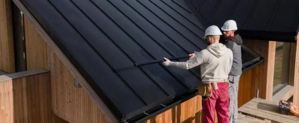 Two construction workers in hard hats installing a black gutter on a dark metal roof of a wooden house.