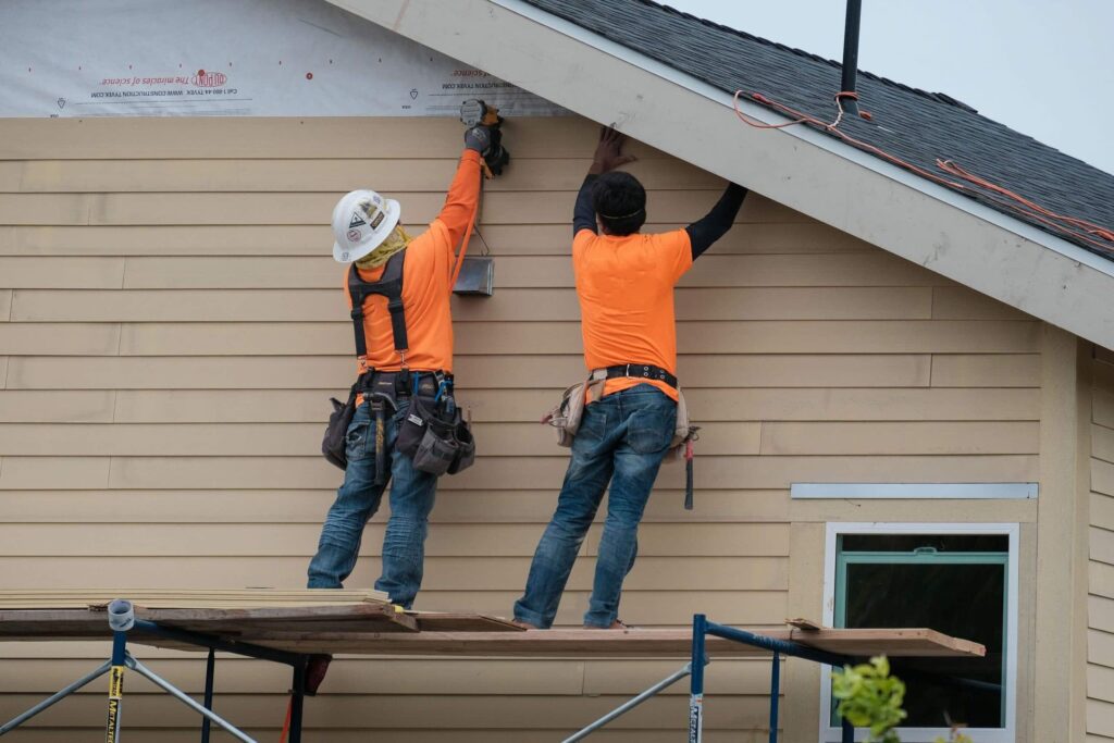 Two construction workers in orange shirts standing on scaffolding, installing beige siding on a house.