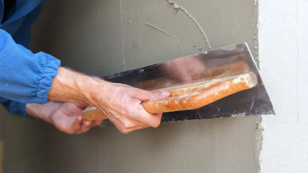 A person using a metal trowel to smooth grey plaster on a wall.