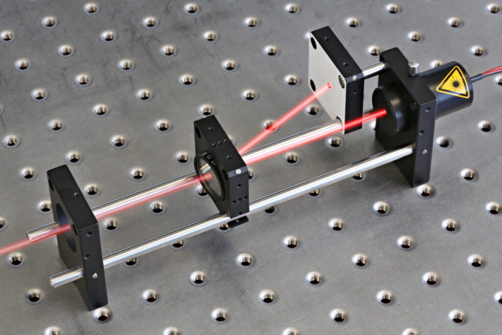 Red laser beam split into two paths by optical mounts on a perforated metal breadboard.