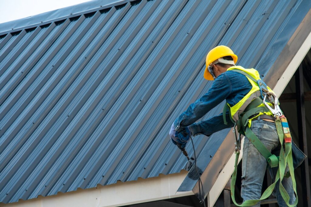 Construction worker in safety gear and a yellow hard hat drilling into a blue corrugated metal roof.
