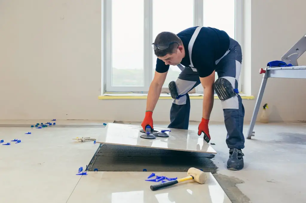 A construction worker in safety gear uses suction cups to lay a large tile onto wet mortar on a floor.