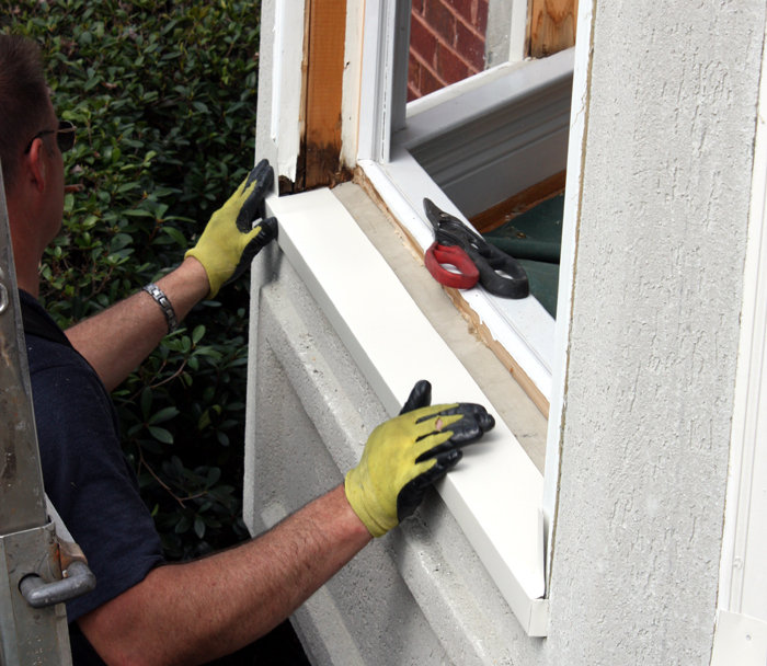 Person wearing yellow work gloves installing white trim around a window on a stucco wall.