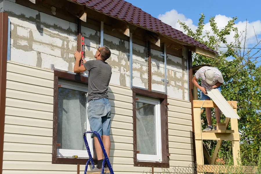 Two men installing beige vinyl siding and brown trim on a house, one using a level on a ladder.