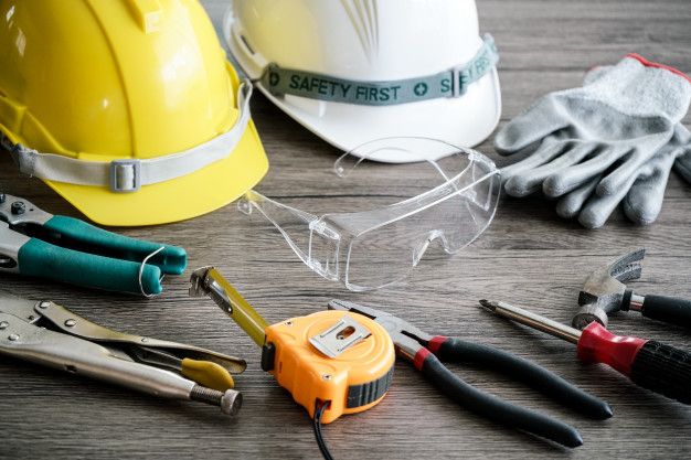 Yellow and white hard hats, safety goggles, work gloves, and hand tools arranged on a wooden background.