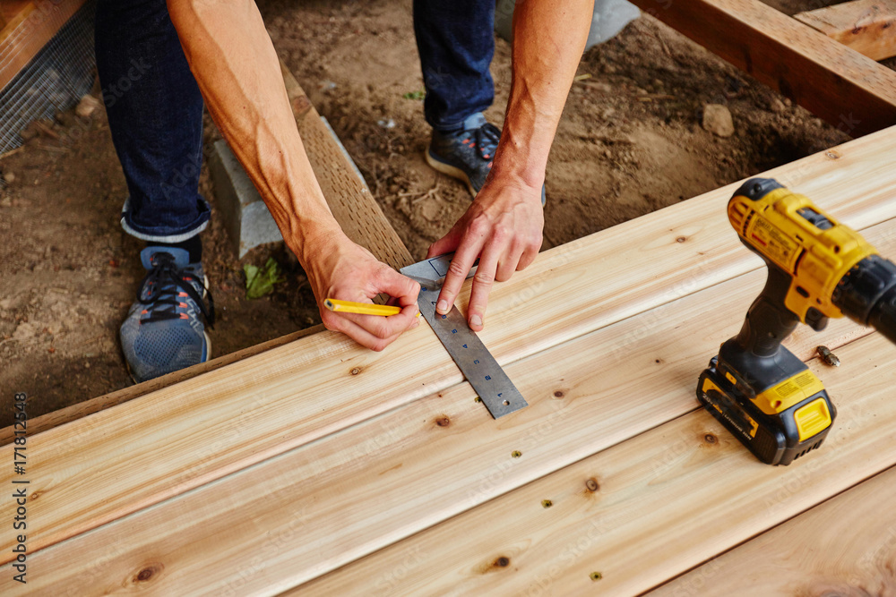 A person uses a carpenter's square and pencil to mark a wooden plank during deck construction.