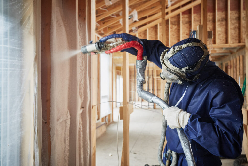 Worker in protective gear spraying foam insulation into wooden wall studs of a house under construction.