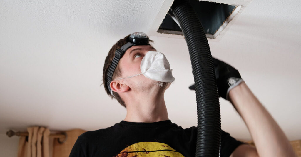 Man wearing a mask and headlamp looking up at a black flexible hose protruding from a ceiling opening.