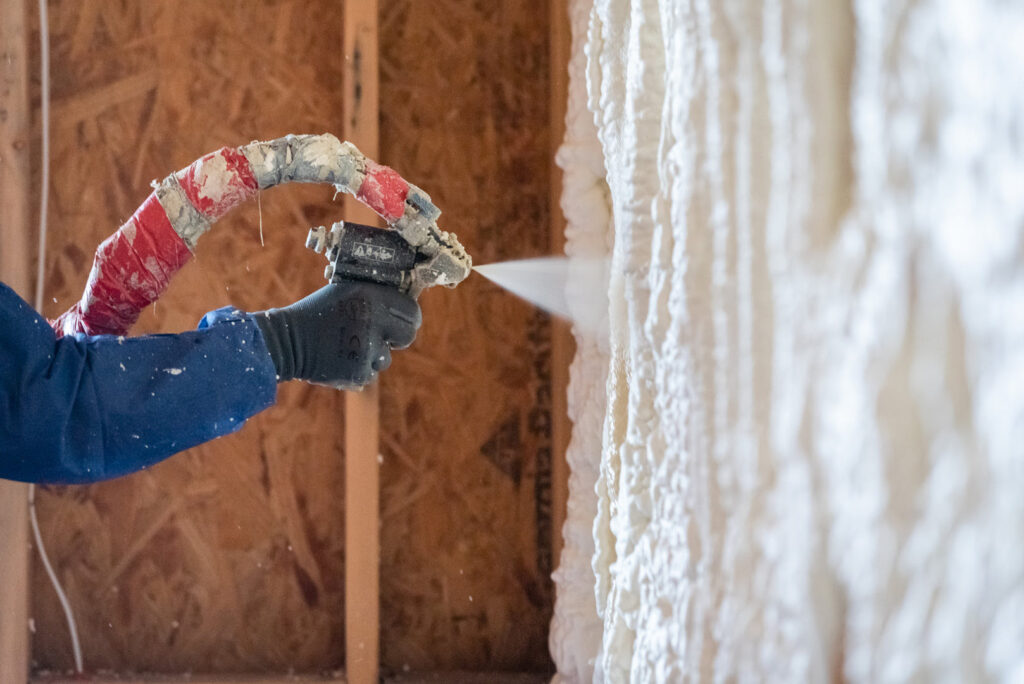 Worker applying white spray foam insulation to a wooden wall using a spray gun.