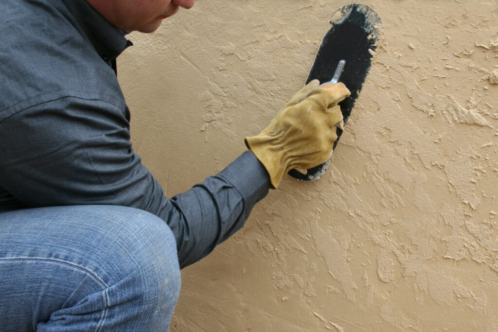 Worker applying tan textured plaster to a wall with a black trowel.