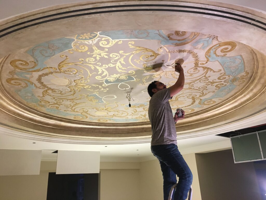 An artist standing on a ladder paints an ornate gold and blue fresco on a domed ceiling.