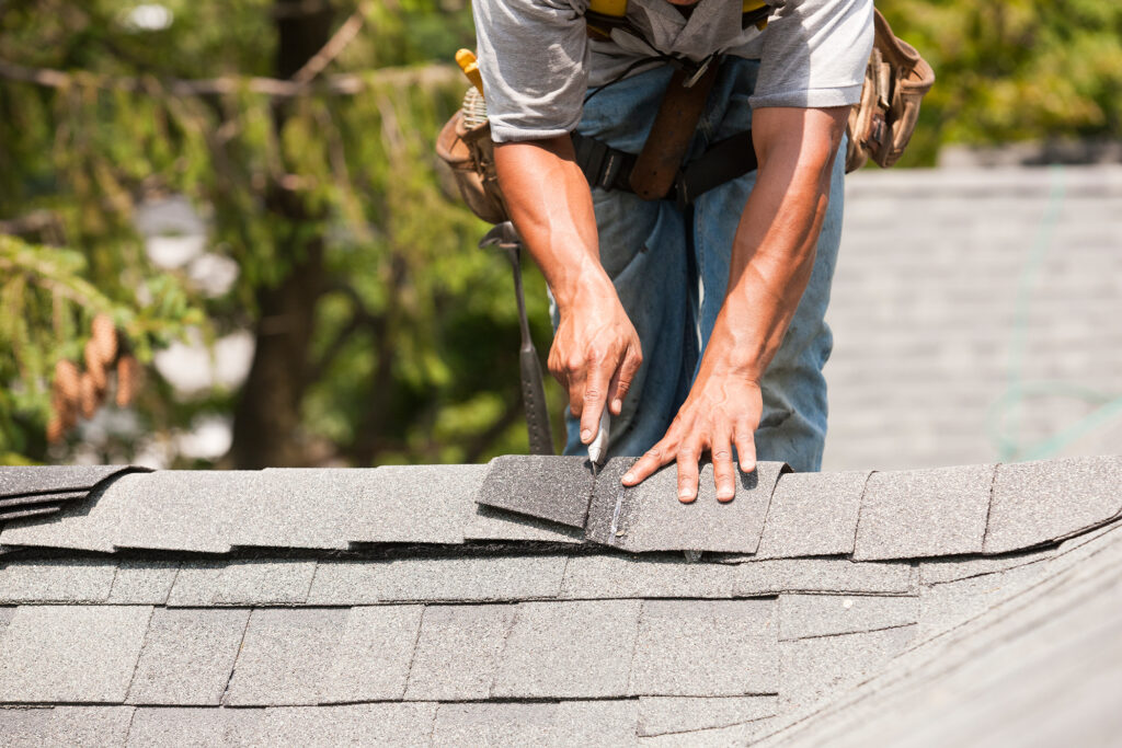 A roofer using a utility knife to cut a grey asphalt shingle on a roof.