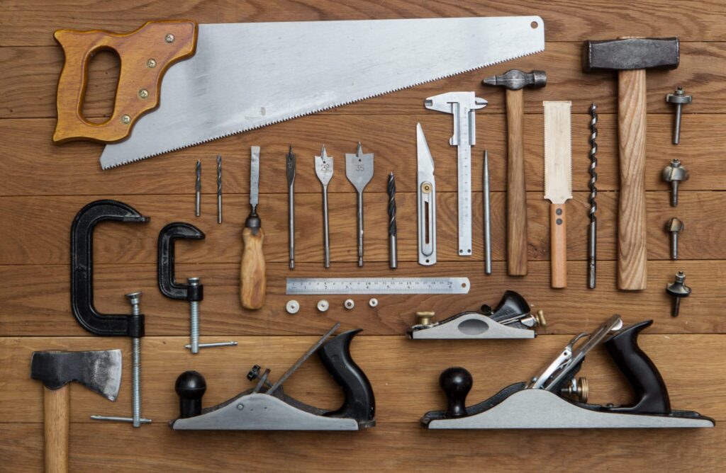 Various woodworking tools, including saws, hammers, planes, clamps, and drill bits, arranged on a wooden surface.