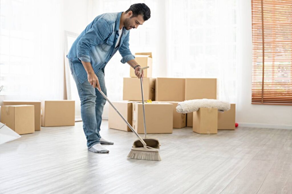 Man sweeping a floor with a broom and dustpan in a room filled with cardboard moving boxes.