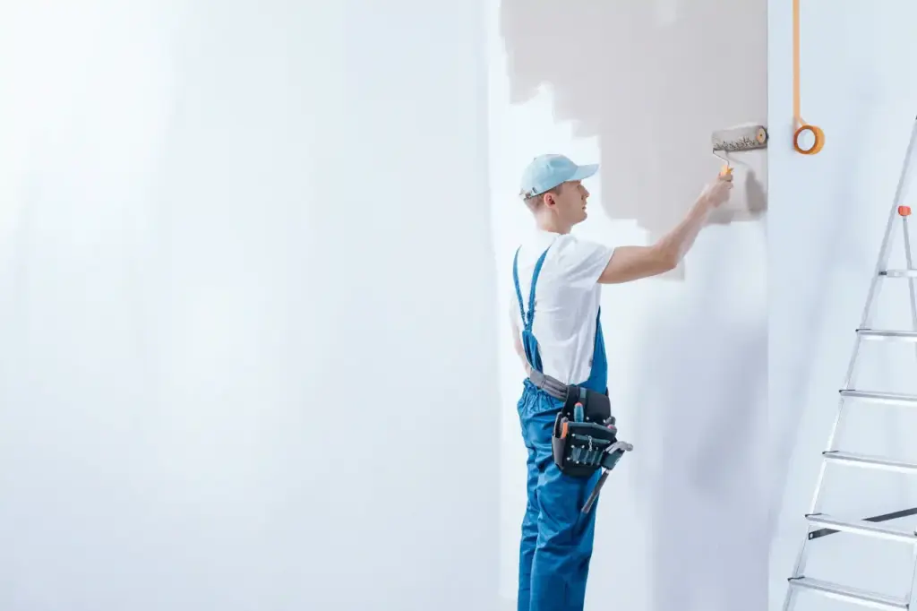 Painter in blue overalls using a paint roller on a white wall.