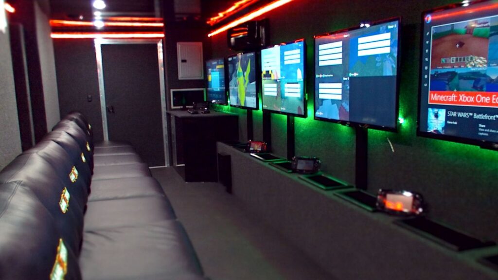 Interior of a mobile gaming trailer with leather bench seating facing a wall of monitors and gaming consoles.