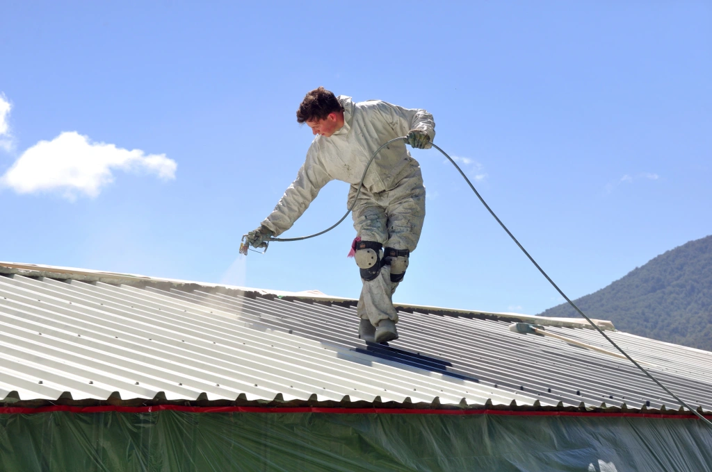 A worker in protective clothing uses a spray gun to apply a coating to a corrugated metal roof against a blue sky.