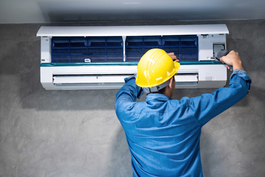 Technician in a yellow hard hat repairing an open wall-mounted air conditioner unit.