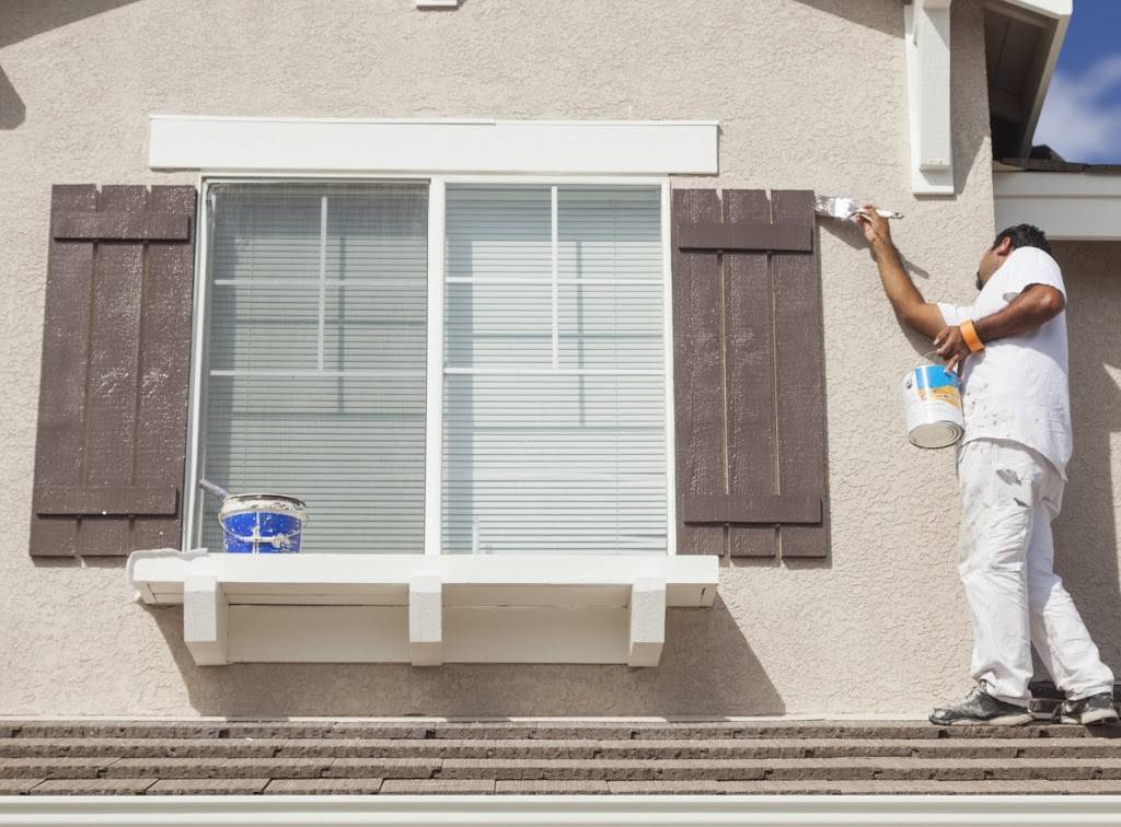 Painter in white work clothes standing on a roof painting brown window shutters.