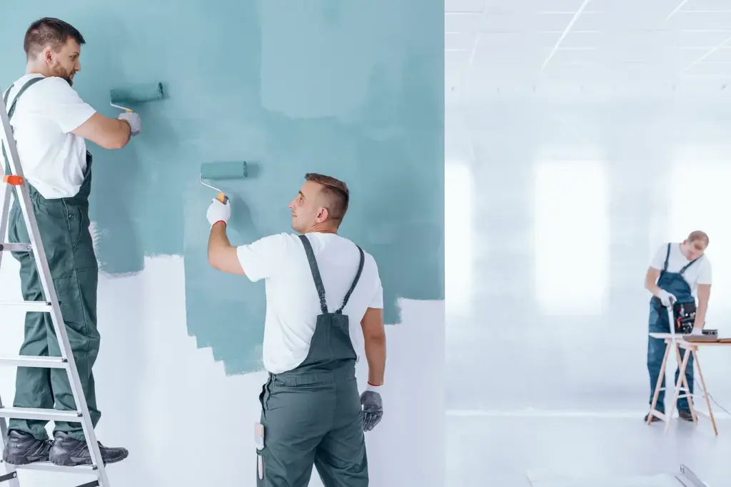 Two painters in overalls paint a wall blue with rollers, while a third worker stands in a bright, white room.