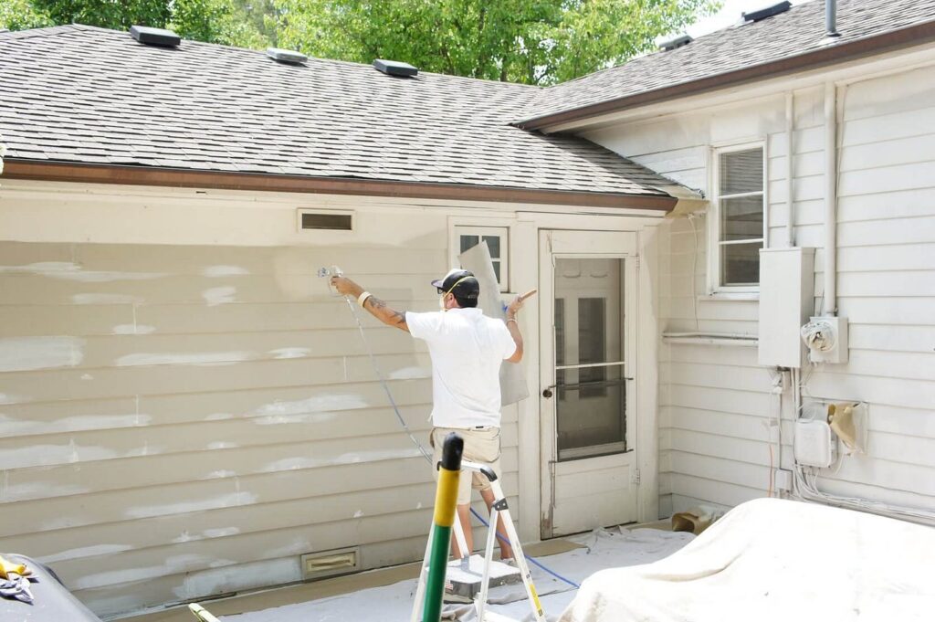 A worker using a paint sprayer to coat the exterior siding of a house.