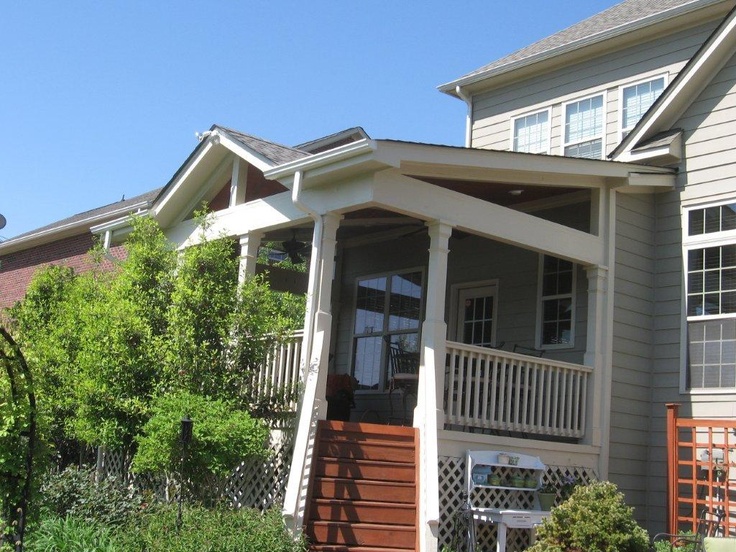Covered back porch with white pillars and wooden stairs attached to a two-story house surrounded by garden foliage.