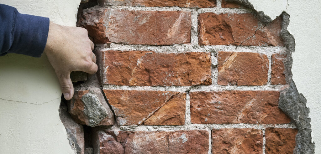 A hand touches an exposed red brick wall where the plaster has cracked and peeled away.
