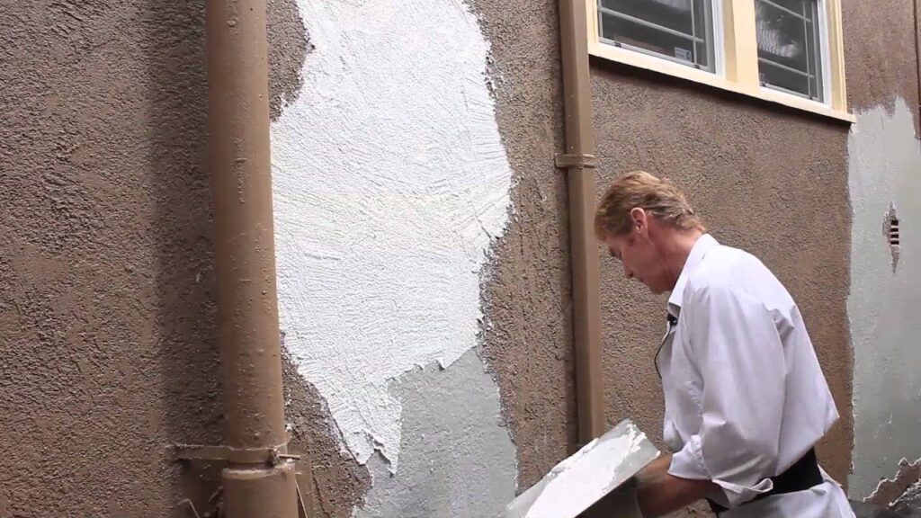 A person applying a white plaster patch to a brown stucco exterior wall.