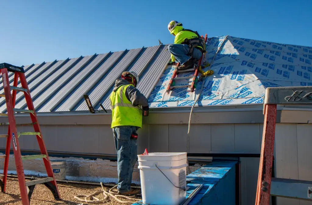Construction workers in high-visibility gear installing a corrugated metal roof.