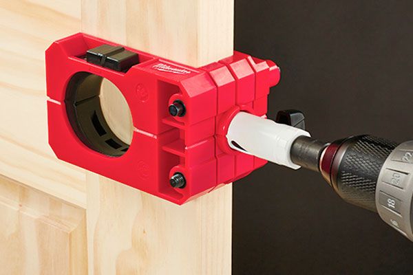 Red drill guide clamped to a wooden beam with a power drill inserted for precise drilling.