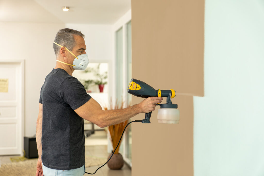 A man in a respirator mask uses a handheld paint sprayer to paint an interior wall beige.