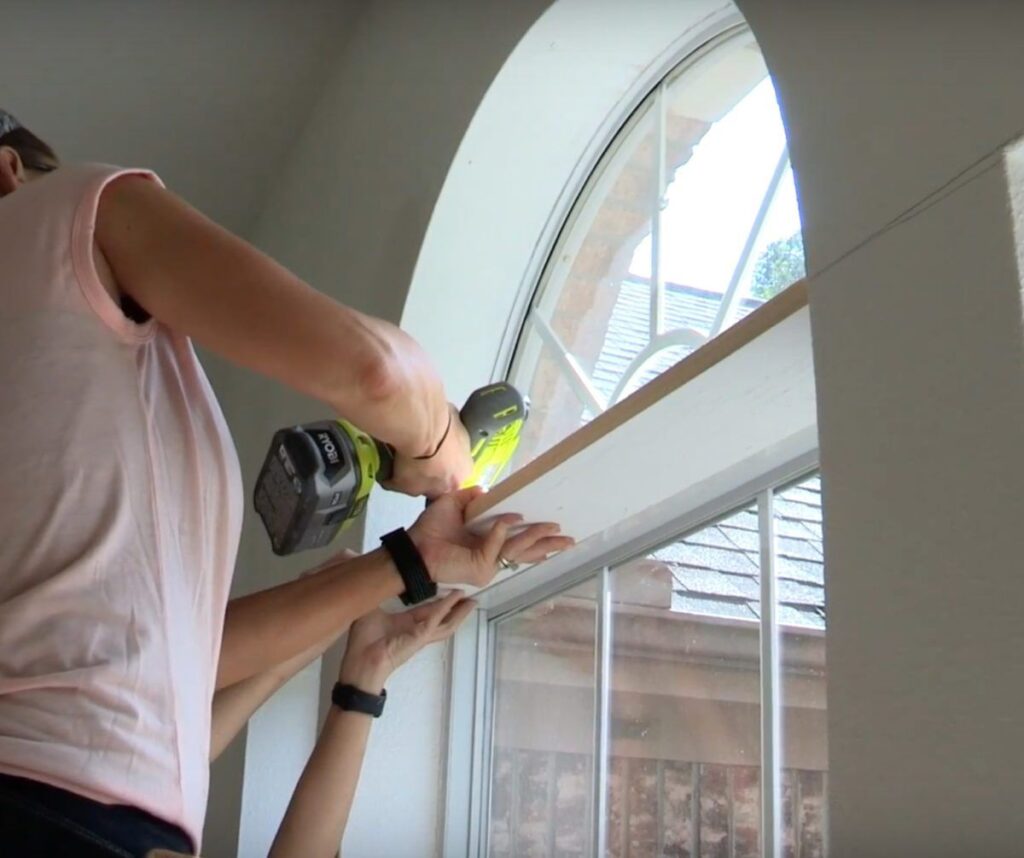 Person installing a wooden board to an arched window frame using a power drill.
