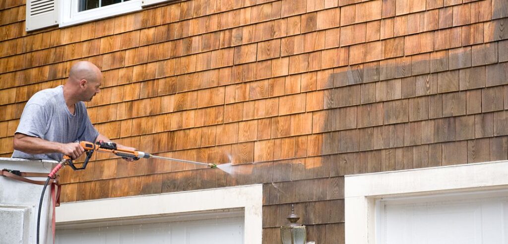 Man using a pressure washer to clean weathered cedar shingles on the side of a house.