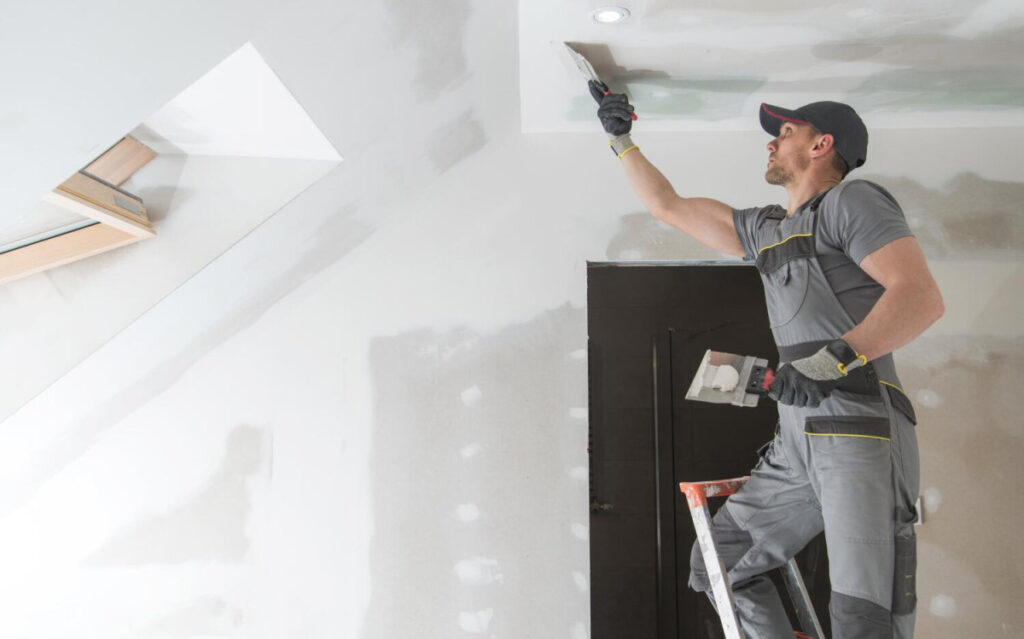 A construction worker on a ladder uses a putty knife to apply plaster to a ceiling in a room under renovation.