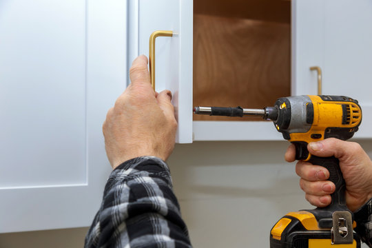 Person using a yellow power drill to install a gold handle on white kitchen cabinets.