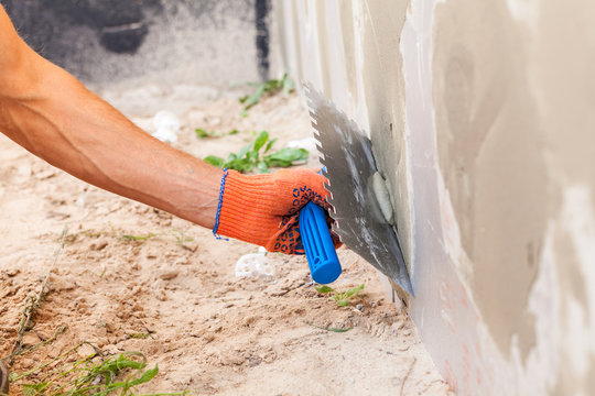 Hand in an orange glove using a notched trowel to apply mortar to an exterior wall.