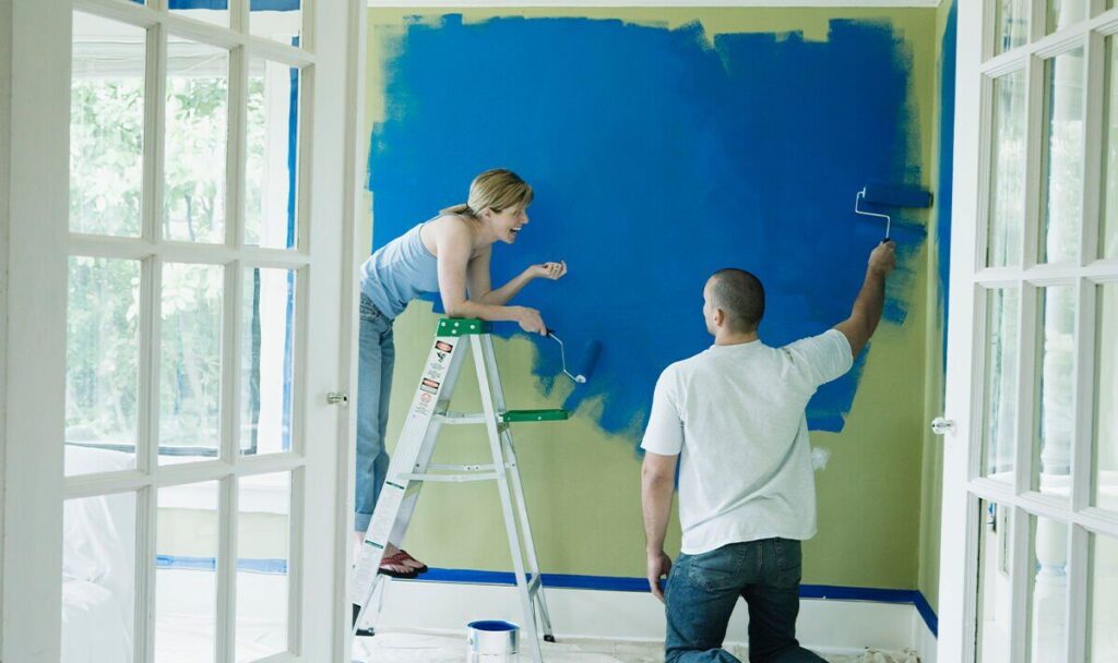 A man and woman happily painting a wall bright blue with paint rollers.