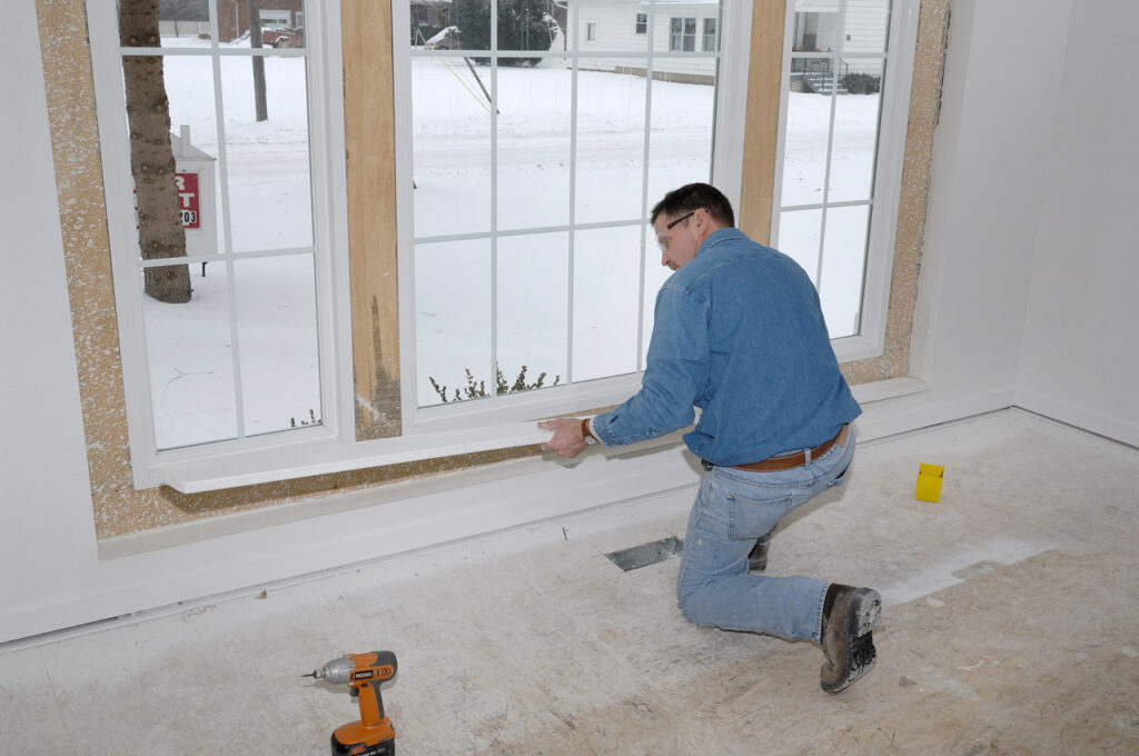 A man in a blue denim shirt kneels while installing white trim around a large window in a house under construction.