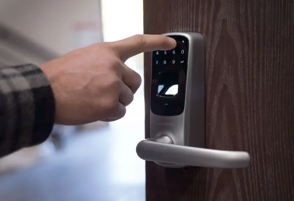 A finger pressing a key on a silver electronic smart lock with a keypad and fingerprint scanner on a wooden door.