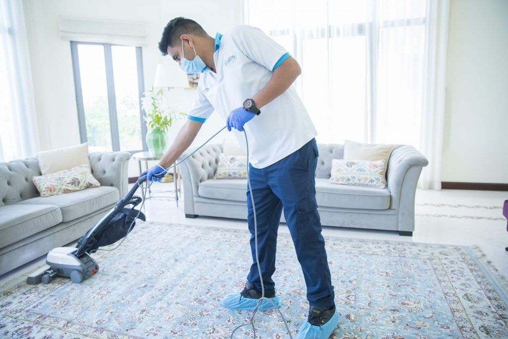 Professional cleaner wearing a mask and gloves vacuuming a patterned carpet in a bright living room.
