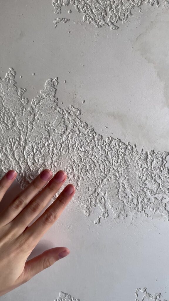 A hand with pink nail polish touches a white wall with patches of peeling, textured paint.