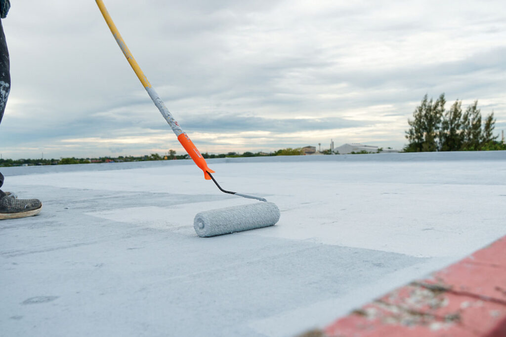 Worker applying white sealant to a flat rooftop with a long-handled paint roller.