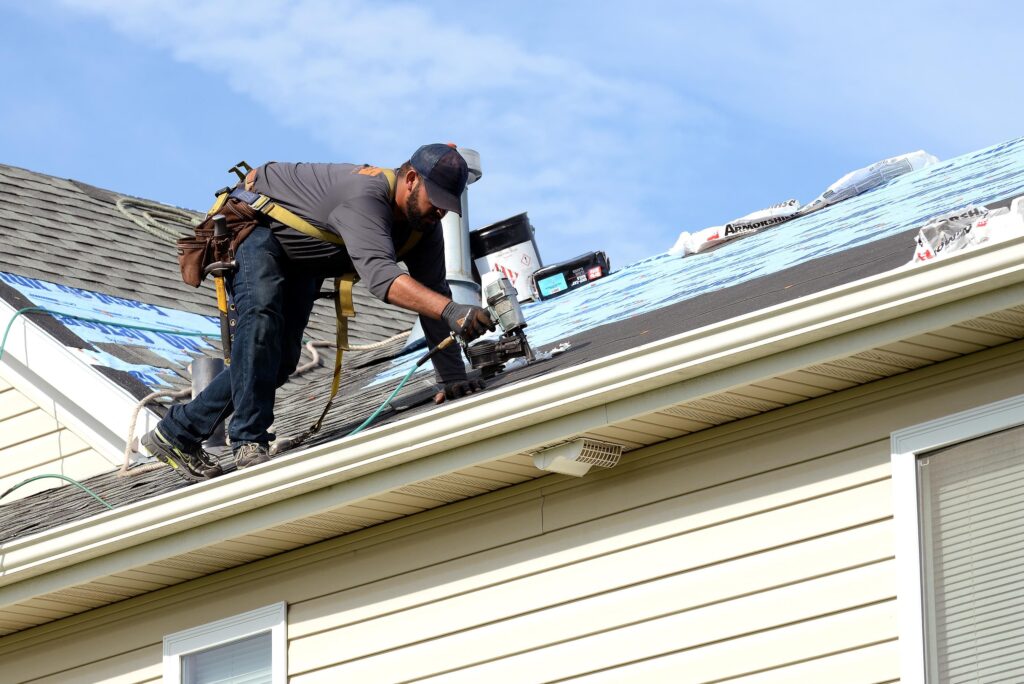 Roofer wearing a safety harness uses a nail gun to install shingles on a house roof.