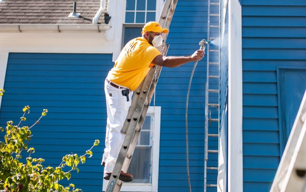A painter in a yellow shirt and mask standing on a ladder while using a spray gun to paint a blue house.