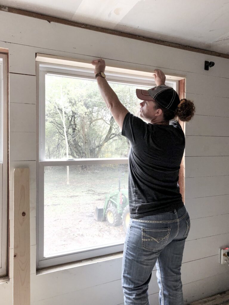 Woman in a baseball cap installing white trim above a window in a room under renovation.