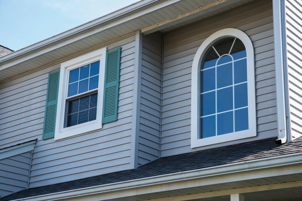 Gray house siding with a rectangular window featuring teal shutters and a nearby arched window.
