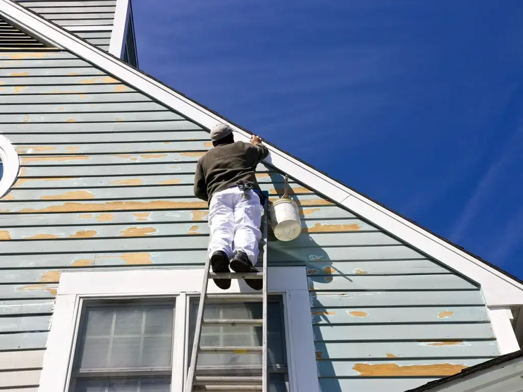 Painter on a ladder scraping the siding of a light blue house in preparation for painting.